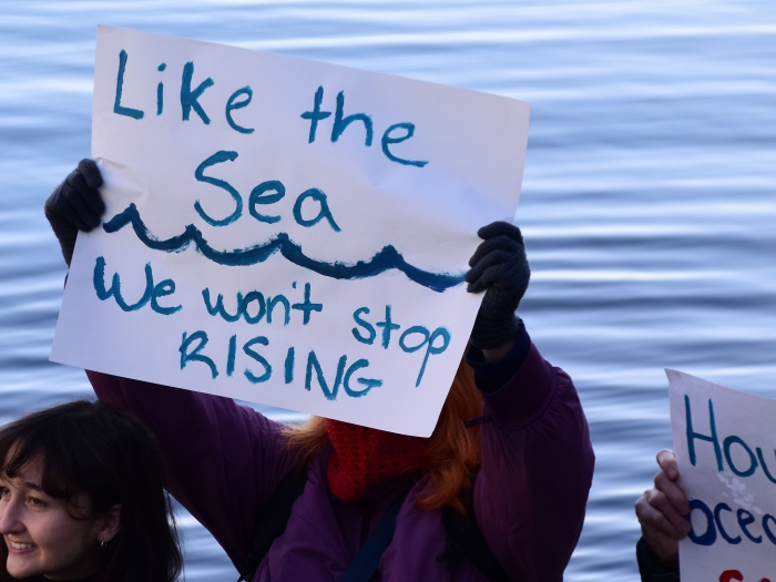 a person at a rally holding a sign that reads "like the sea, we won't stop rising"
